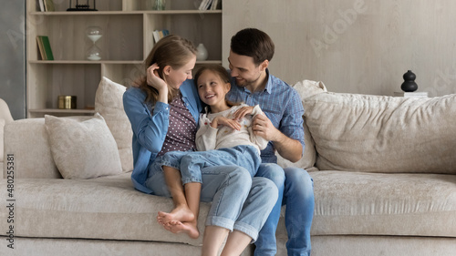 Loving caring happy young couple parents tickling adorable little preteen kid daughter, having fun sitting on cozy couch together in modern living room, enjoying carefree playtime weekend activity.