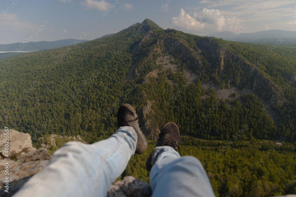 A man sits with his legs dangling in the mountains against the backdrop of a large mountain.