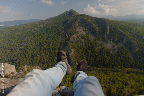 A man sits with his legs dangling in the mountains against the backdrop of a large mountain.