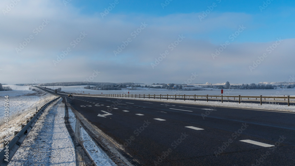 Fototapeta premium Country road through winter rural scene. Black clean asphalt. Winter road going through snow-covered fields.