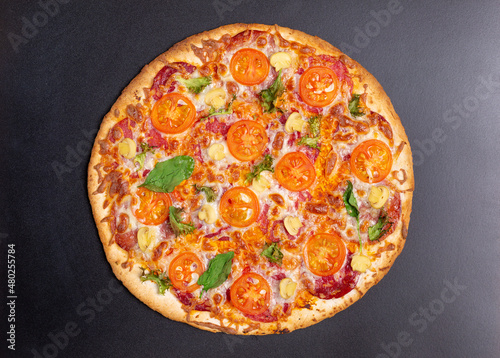 Freshly baked pizza with several types of sausage, ham, cherry tomatoes, mushrooms, basil leaves and lettuce on a black background.