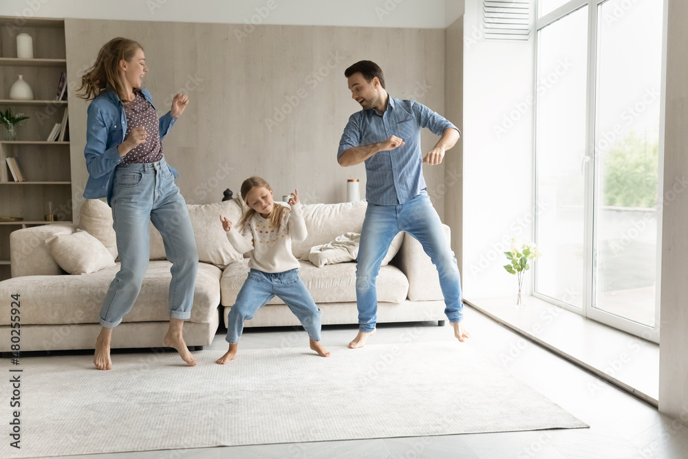 Overjoyed sincere millennial couple parents and small adorable laughing child daughter listening energetic disco music, making funny body moves, entertaining together at modern home on weekend.