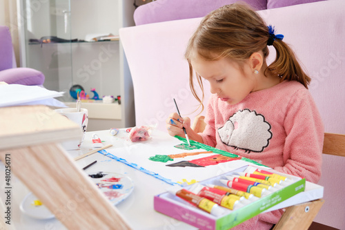 Little girl sits by desk and paints a picture with acrylic paints.