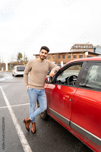 Young latin or arab eastern indian pakistani carefree man in stylish clothes standing near red small car on parking slot of an urban city background on rainy midseason day. Travel, rental, car sharing © pavelgulea