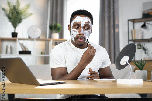 Cosmetic face mask procedures where young handsome african american applying white clay mask on his face with brush. Beauty and skin treatment procedures
