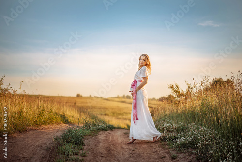 A pregnant woman in a white dress stands barefoot on a path in a field on a sunny summer day.