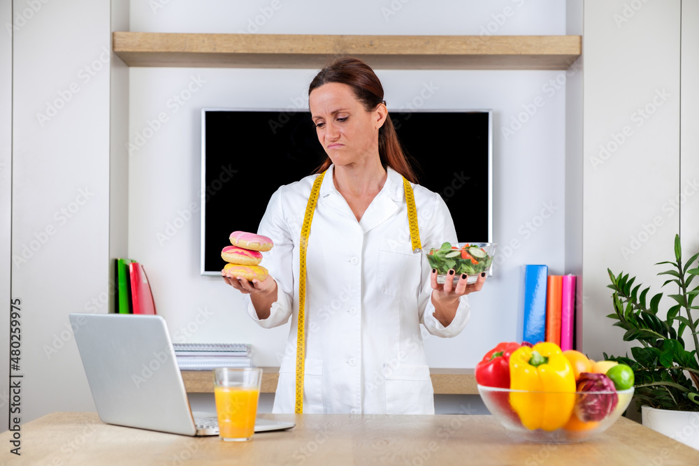 Young woman wearing nutritionist uniform holding a salad and donuts