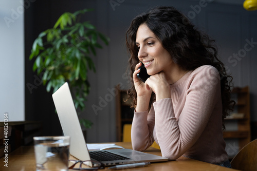 Excited businesswoman or freelance consultant talk on cell sitting at desk with laptop. Young woman remote advisor consult client by phone call negotiating, discuss project or contract with colleague photo