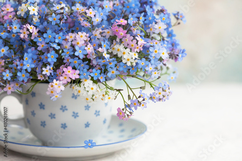 Fototapeta Naklejka Na Ścianę i Meble -  Bouquet of spring forget-me-not flowers in a cup on the table against the background of a colored wall. Holiday card, fragment, blur, selective focus.