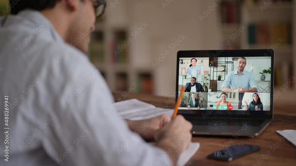 Video call, online conference using app and laptop. Over shoulder view of student or freelancer, studies or works remotely, watches an online lecture, takes notes, multiracial people on laptop screen Video call, online conference using app and laptop. Over shoulder view of student or freelancer, studies or works remotely, watches an online lecture, takes notes, multiracial people on laptop screen