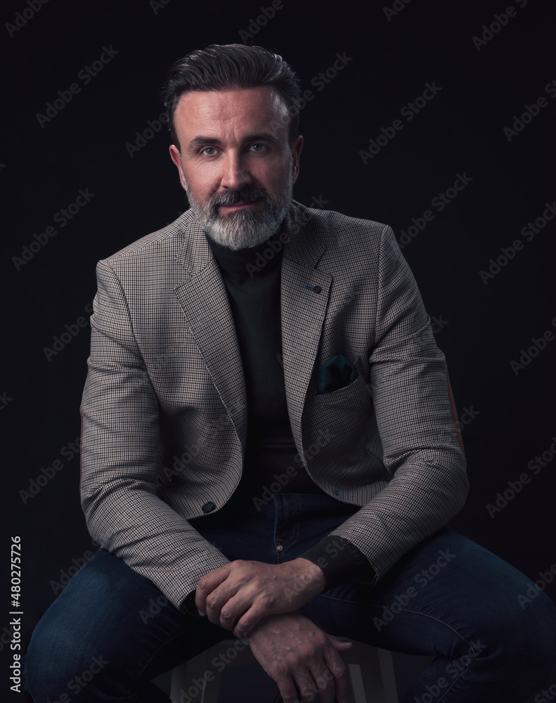 Portrait of adult businessman wearing trendy suit and sitting in modern studio on stylish chair against the black background Portrait of adult businessman wearing trendy suit and sitting in modern studio on stylish chair against the black background