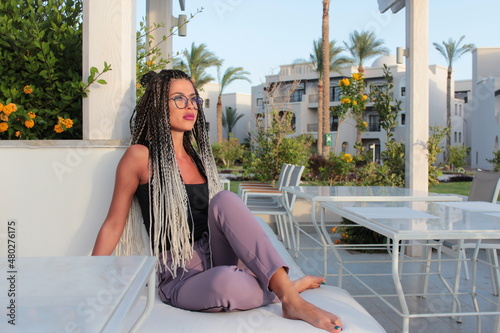 Foto A woman with African braids, a sultry, sexy beauty in glasses and in a suit with plump lips, sits on a couch outdoors around her palm trees