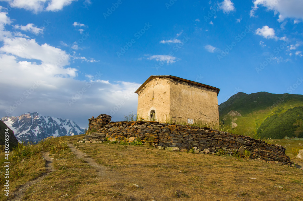 blue sky with clouds, green grass and caucasus mountains around the  ancient stone church in Svaneti, Georgia