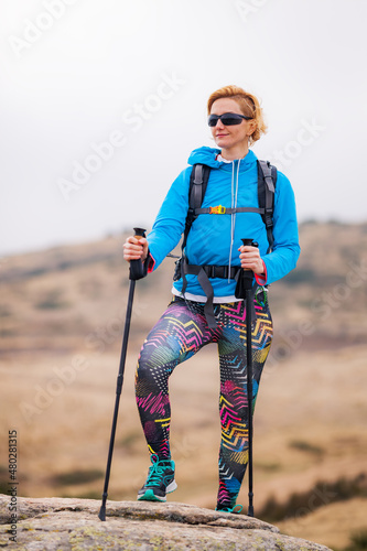 Hiking girl with poles and backpack standing on rocks. Windy autumn day. Travel and healthy lifestyle outdoors in fall season
