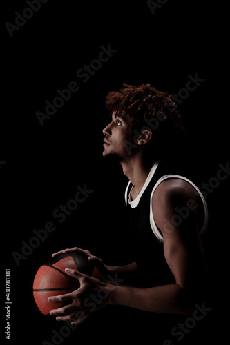 Professional basketball player holding a ball against black background. Serious concentrated african american man in sports uniform © Nikola Spasenoski