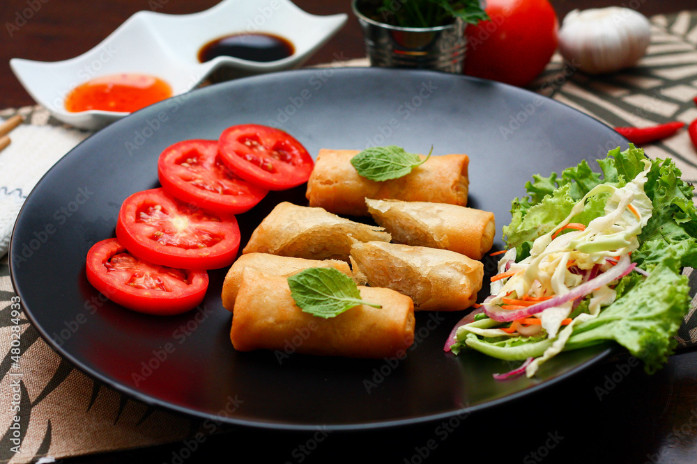 Fried spring rolls with vegetables and tomatoes placed in a black plate on a black wooden table and dipping sauce. Fried spring rolls with vegetables and tomatoes placed in a black plate on a black wooden table and dipping sauce.
