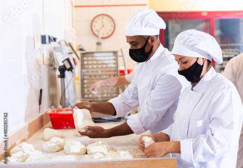 Young Latina working with husband in small family bakery, shaping dough on floured surface before baking