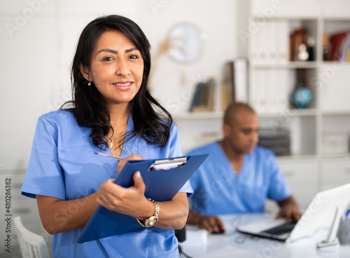 Polite latina female health worker meeting patient in medical office, filling out medical form at clipboard.