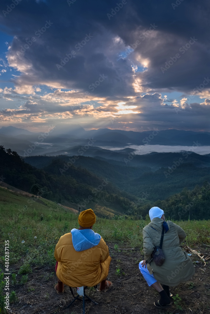 Two man is on a top of hill with sea of fog, mountain range silhouette and colorful light beam in a sunrise morning sky 