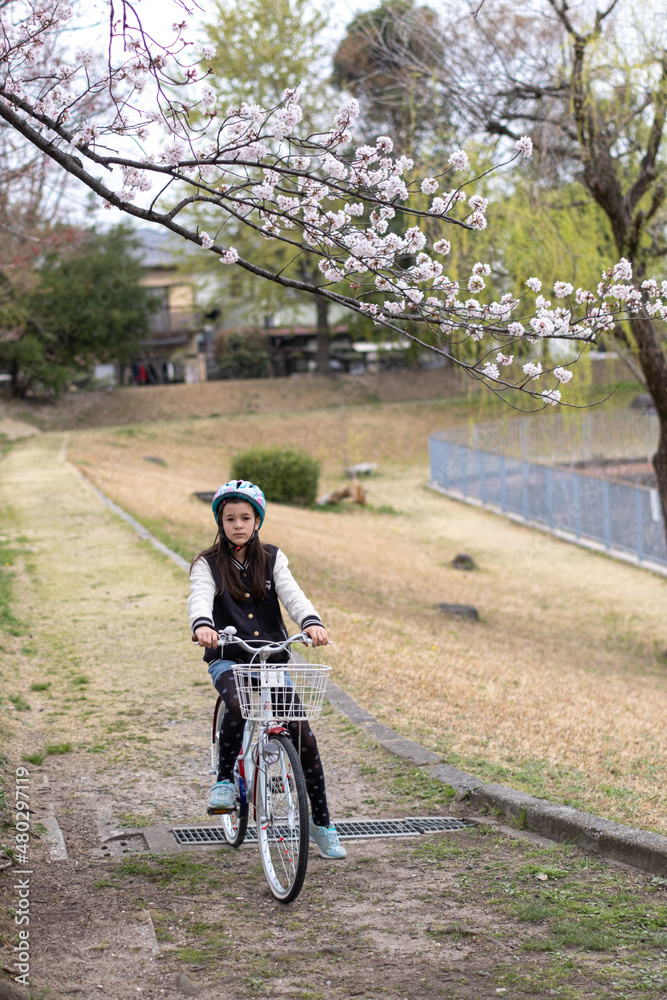Preteen girl on a bicycle, standing under a blooming branch of sakura cherry blossom tree with a sad face. Springtime.
