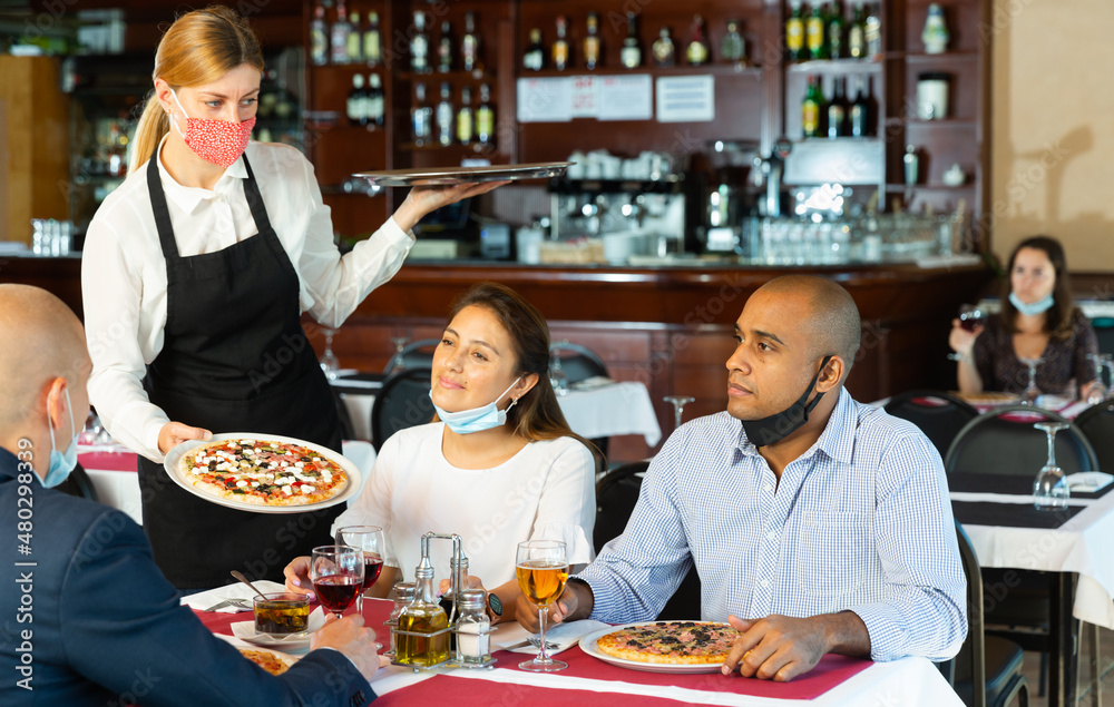 Young waitress in protective face mask serving pizza to friendly company in restaurant. Precautions in catering establishments during pandemic coronavirus