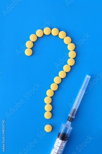 A question symbol is lined with yellow pills on a blue background and a syringe lies nearby.