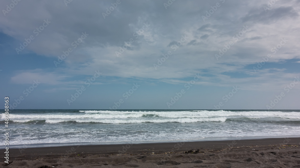 Pacific Ocean coast. Powerful waves with white foam roll onto the shore with volcanic black sand. Clouds in the blue sky. Kamchatka. Halaktyrsky beach