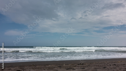 Pacific Ocean coast. Powerful waves with white foam roll onto the shore with volcanic black sand. Clouds in the blue sky. Kamchatka. Halaktyrsky beach
