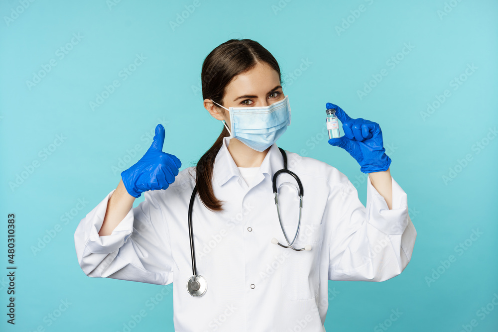 Portrait of smiling doctor, medical personel in face medical mask and rubber gloves, showing thumbs up and coronavirus, omicron vaccine, standing over blue background - obrazy, fototapety, plakaty 