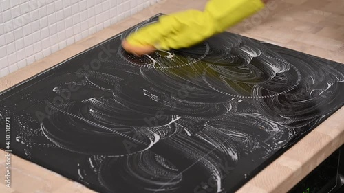 Cleaning cooktop cooking panel in kitchen with fat remover spray and blue rag by a woman in yellow rubber gloves.