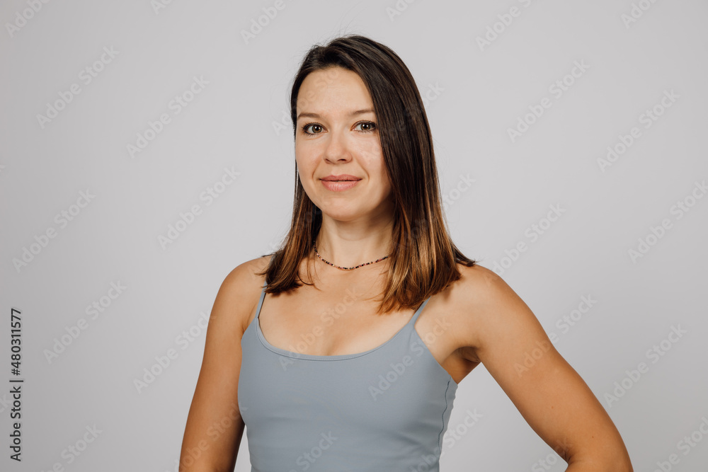 Portrait of a smiling brunette. Beautiful happy young woman in gray top for yoga or fitness poses on gray isolated background in studio, she is standing sideways. Portrait of a smiling brunette. Beautiful happy young woman in gray top for yoga or fitness poses on gray isolated background in studio, she is standing sideways.