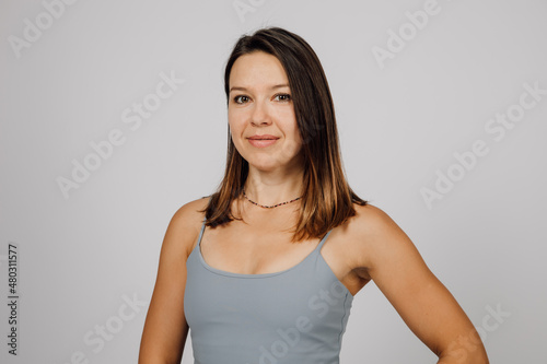 Portrait of a smiling brunette. Beautiful happy young woman in gray top for yoga or fitness poses on gray isolated background in studio, she is standing sideways.