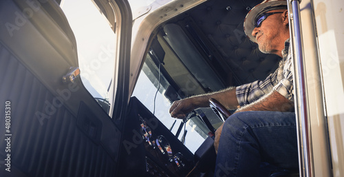 Elderly Latino semi truck driver sits in the cab looking ahead wearing plaid shirt, sunglasses and hard hat. Handsome cool looking trucker sits at the steering wheel.  