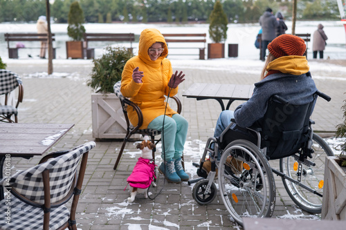 Two girlfriends in a cafe on a street terrace in winter. Woman in a wheelchair.