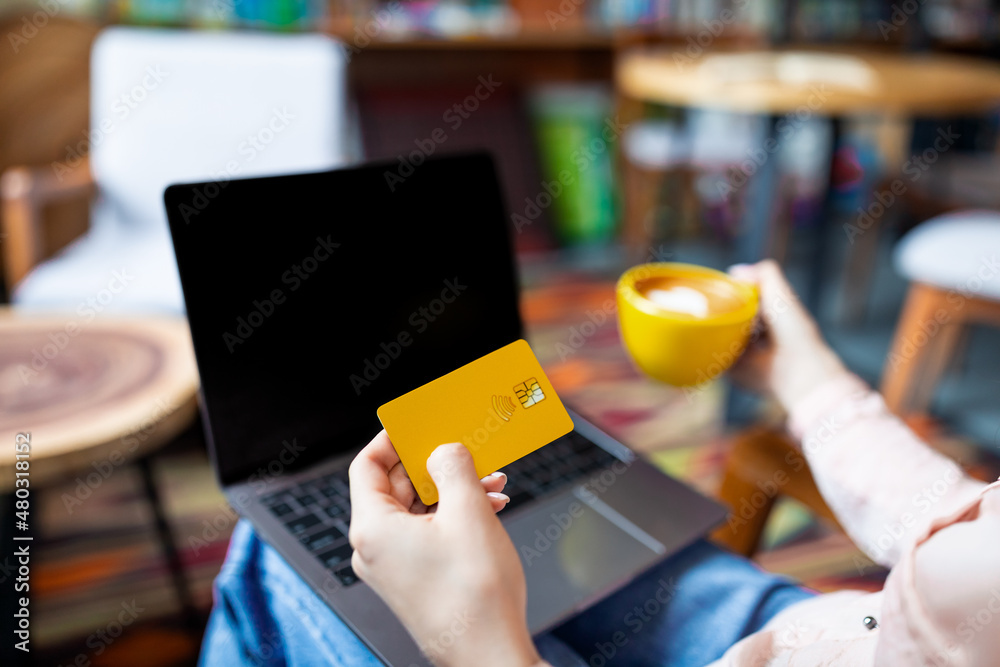 Contactless payment. Woman holding credit card and drinking coffee while sitting in cafeteria and working on laptop Contactless payment. Woman holding credit card and drinking coffee while sitting in cafeteria and working on laptop