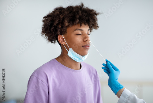 Doctor in rubber gloves making nasal coronavirus PCR test for African American teen, using sterile swab stick indoors