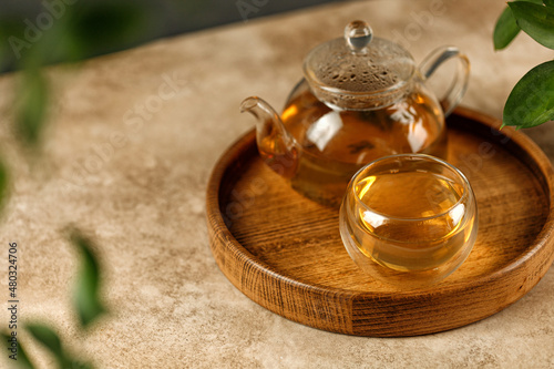 Double Wall Glass Cup with green herbal tea and glass teapot on wood tray and beige background photo