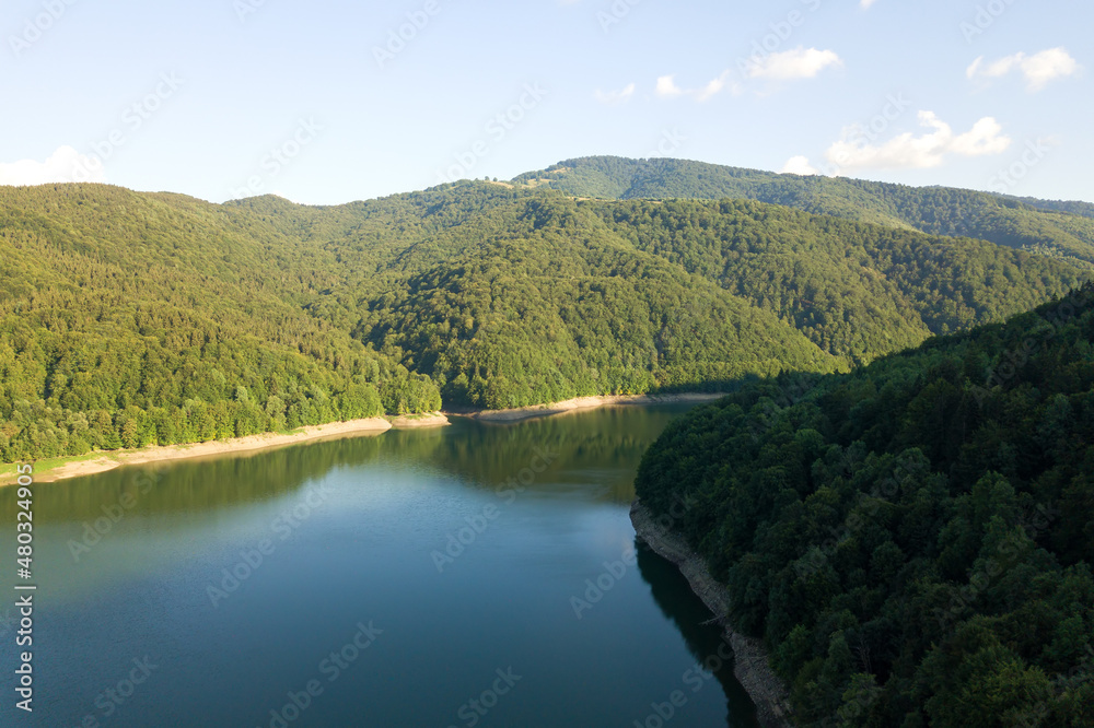 Aerial view of big lake with clear blue water between high mountain hills covered with dense evergreen forest.