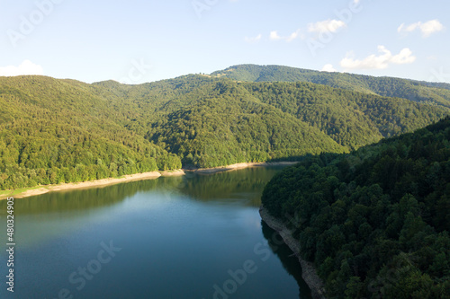 Aerial view of big lake with clear blue water between high mountain hills covered with dense evergreen forest.