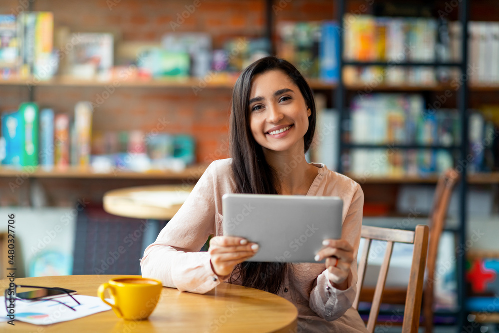 Happy arab woman sitting at cafe, using digital tablet and drinking coffee, smiling at camera, copy space