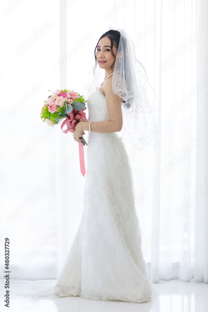 Studio shot of Asian young happy beautiful luxury long hair female bride in white wedding dress standing smiling posing holding pink roses flower bouquet in front of curtain in engagement ceremony Studio shot of Asian young happy beautiful luxury long hair female bride in white wedding dress standing smiling posing holding pink roses flower bouquet in front of curtain in engagement ceremony