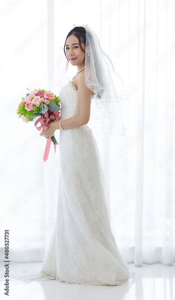 Studio shot of Asian young happy beautiful luxury long hair female bride in white wedding dress standing smiling posing holding pink roses flower bouquet in front of curtain in engagement ceremony