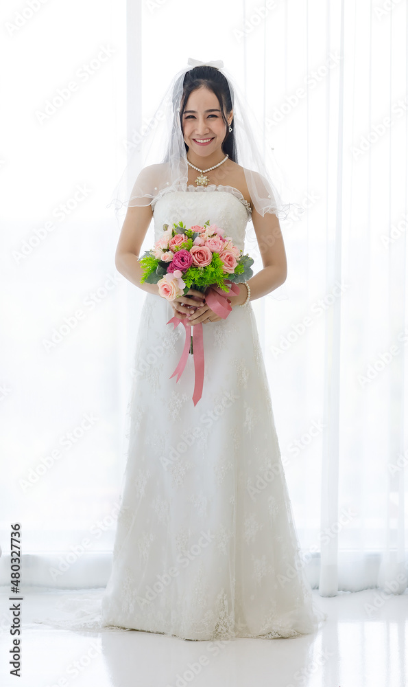 Studio shot of Asian young happy beautiful luxury long hair female bride in white wedding dress standing smiling posing holding pink roses flower bouquet in front of curtain in engagement ceremony Studio shot of Asian young happy beautiful luxury long hair female bride in white wedding dress standing smiling posing holding pink roses flower bouquet in front of curtain in engagement ceremony