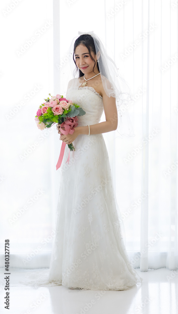 Studio shot of Asian young happy beautiful luxury long hair female bride in white wedding dress standing smiling posing holding pink roses flower bouquet in front of curtain in engagement ceremony Studio shot of Asian young happy beautiful luxury long hair female bride in white wedding dress standing smiling posing holding pink roses flower bouquet in front of curtain in engagement ceremony