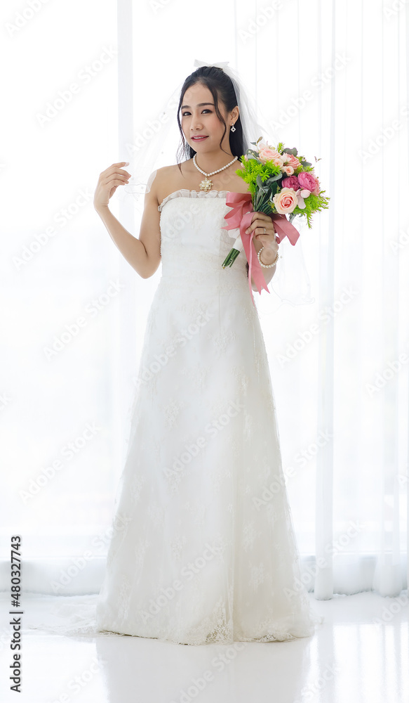 Studio shot of Asian young happy beautiful luxury long hair female bride in white wedding dress standing smiling posing holding pink roses flower bouquet in front of curtain in engagement ceremony Studio shot of Asian young happy beautiful luxury long hair female bride in white wedding dress standing smiling posing holding pink roses flower bouquet in front of curtain in engagement ceremony