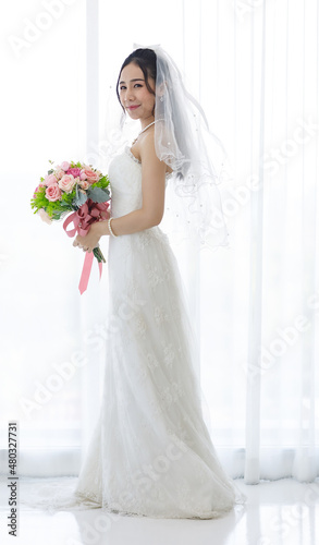 Studio shot of Asian young happy beautiful luxury long hair female bride in white wedding dress standing smiling posing holding pink roses flower bouquet in front of curtain in engagement ceremony