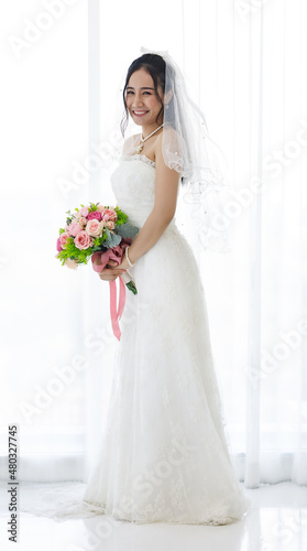 Studio shot of Asian young happy beautiful luxury long hair female bride in white wedding dress standing smiling posing holding pink roses flower bouquet in front of curtain in engagement ceremony