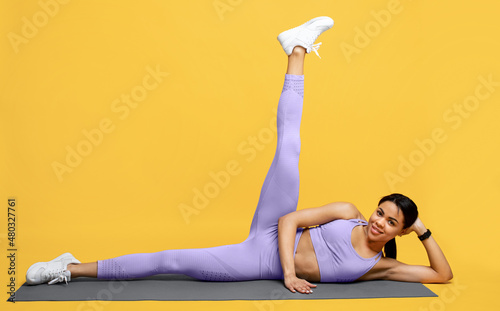 Fototapeta Naklejka Na Ścianę i Meble -  Young sporty black lady warming up and lifting leg up, exercising on mat over yellow background