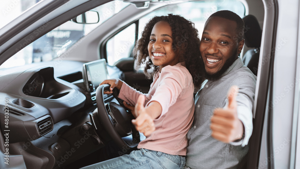 Happy black father and daughter sitting on driver seat of new car, showing thumb up, recommending auto dealership