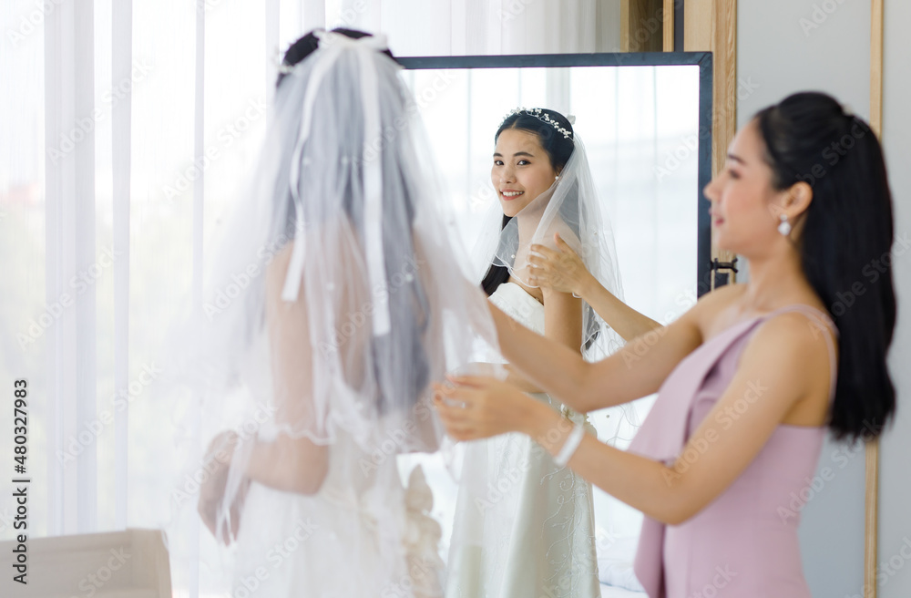 Naklejka premium Asian lovely beautiful happy cheerful female bride in white long wedding dress standing staring at her reflection in mirror while pretty bridesmaid helping adjusting facial veil in blurred foreground